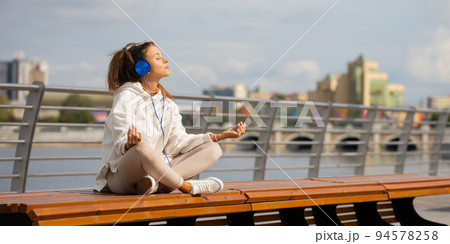 Young woman meditates on the embankment - she sitting in headphones and listens to relaxing music. 94578258
