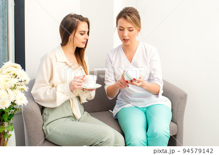 Beautician offering product for the young woman holding a white plastic jar with a cream sitting on the sofa 94579142