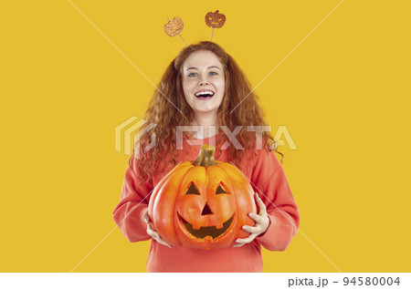 Portrait of cheerful young woman holding Halloween pumpkin with funny carved face on it. 94580004