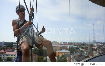 Industrial mountaineering worker in protective helmet showing approval gesture and smiling while hanging on rope behind window. Man using safety lifting equipment outside building. Industrial mountaineering worker in protective helmet showing approval gesture and smiling while hanging on rope behind window. Man using safety lifting equipment outside building. 94583385