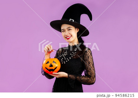 Halloween theme, young asian pretty woman in witch costume wearing black witch hat holding pumpkin bucket posing on purple color background. 94586215