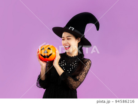 Halloween theme, happy asian woman in elegrant black dress wearing witch hat holding evil orange pumpkin on purple color background.. 94586216