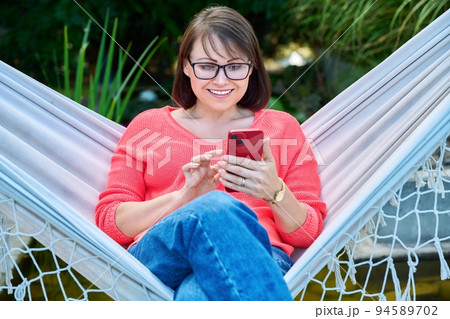 Middle aged woman with smartphone sitting in hammock 94589702