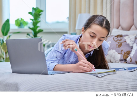Teenage female student lies on bed with laptop, studying remotely Teenage female student lies on bed with laptop, studying remotely 94589773