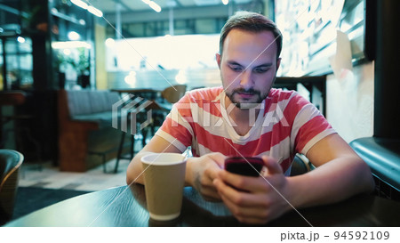 Young attractive man with a beard uses a smartphone in a cafe. Young attractive man with a beard uses a smartphone in a cafe. 94592109