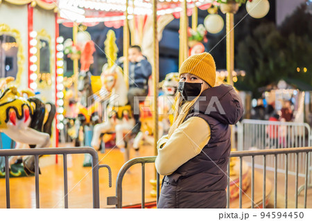 Girl put on a medical mask against coronavirus COVID 19 walking in Christmas traditional market decorated with holiday lights in the evening. Winter portrait beautiful young woman outdoors in front of Girl put on a medical mask against coronavirus COVID 19 walking in Christmas traditional market decorated with holiday lights in the evening. Winter portrait beautiful young woman outdoors in front of 94594450