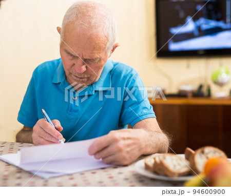 Focused senior man reading and signing papers at home table 94600900