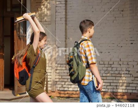 schoolchildren are having fun in the school yard - a teenage girl wants to hit a boy on the head with books 94602118