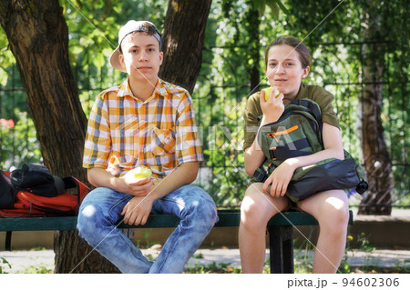portrait of students in a city park, teenage schoolchildren a boy and a girl sitting on a bench, resting and eating apples portrait of students in a city park, teenage schoolchildren a boy and a girl sitting on a bench, resting and eating apples 94602306