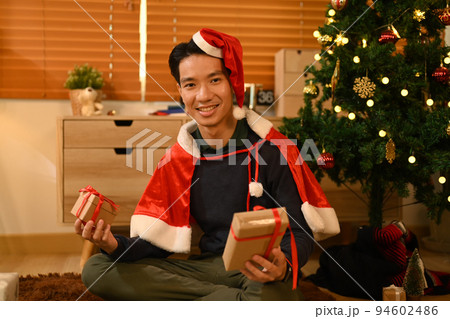 Handsome man in Santa hat holding Christmas gifts and smiling to camera, celebrating New year and Christmas festive Handsome man in Santa hat holding Christmas gifts and smiling to camera, celebrating New year and Christmas festive 94602486