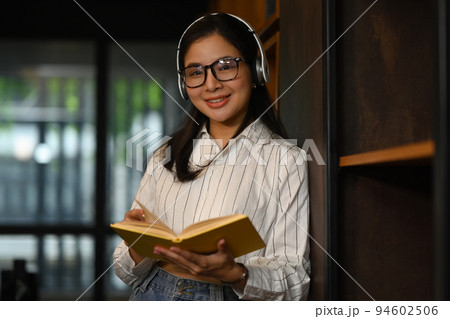 Positive female student reading a book between library bookshelves. Education and people concept 94602506