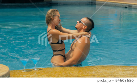 The loving couple hugs and kisses, drinking blue cocktail alcohol liquor in swimming pool at hotel outdoor. Portrait of caucasian man and woman. 94603680