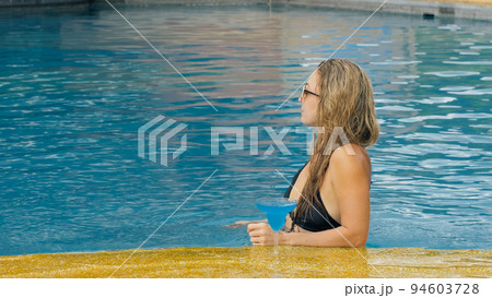 The wide middle shot pretty woman drinking blue cocktail alcohol liquor in swimming pool at hotel. Portrait of sexy girl in pool outdoor. 94603728