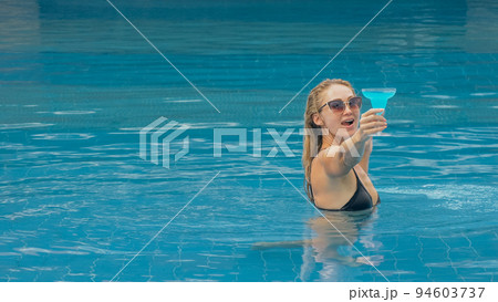The wide middle shot pretty woman drinking blue cocktail alcohol liquor in swimming pool at hotel. Portrait of sexy girl in pool outdoor. 94603737