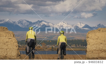 The man and woman travel on mixed terrain cycle touring with bikepacking. The two people journey with bicycle bags. Mountain snow capped, stone arch. The man and woman travel on mixed terrain cycle touring with bikepacking. The two people journey with bicycle bags. Mountain snow capped, stone arch. 94603832