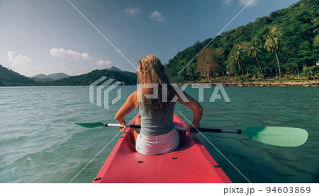Long haired blonde woman with sunglasses rows bright pink canoe along sea bay water to beach with growing palms. Long haired blonde woman with sunglasses rows bright pink canoe along sea bay water to beach with growing palms. 94603869