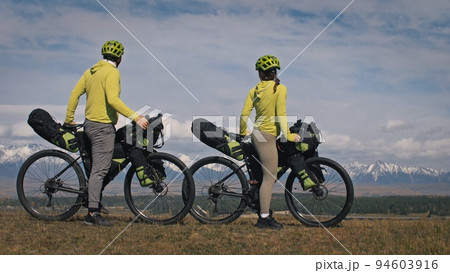 The man and woman travel on mixed terrain cycle touring with bikepacking. The two people journey with bicycle bags. Mountain snow capped. The man and woman travel on mixed terrain cycle touring with bikepacking. The two people journey with bicycle bags. Mountain snow capped. 94603916