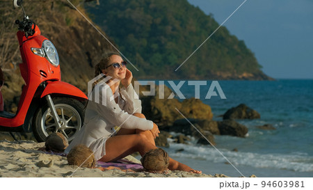 Scooter road trip. Woman alone on red motorbike in white clothes on sand beach by ocean. One girl caucasian tourist walk near tropical palm tree. Scooter road trip. Woman alone on red motorbike in white clothes on sand beach by ocean. One girl caucasian tourist walk near tropical palm tree. 94603981