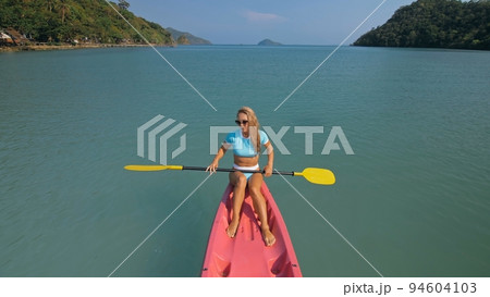 Pretty sportive blonde lady in stylish swimsuit sails on pink plastic canoe along azure ocean bay at exotic resort. 94604103