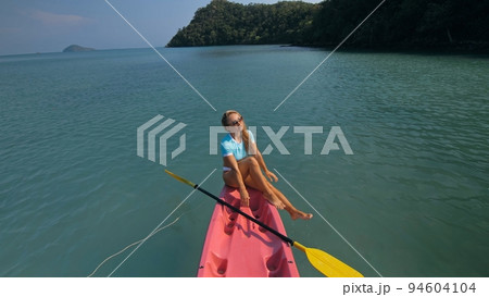Pretty sportive blonde lady in stylish swimsuit sails on pink plastic canoe along azure ocean bay at exotic resort. 94604104