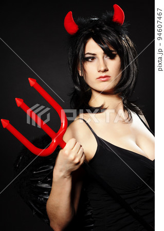 A beautiful, sexy woman in a devil costume, a demon with horns and pitchforks, a trident, in a red dress. American, dress for a costume party Halloween. Choker on the neck. Bright makeup 94607467