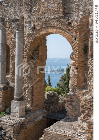 Columns and arch at old ruins of ancient Greek theater at coastal city. Historic building structure at Giardini Naxos bay with sea in background. View of famous tourist attraction on sunny day. Columns and arch at old ruins of ancient Greek theater at coastal city. Historic building structure at Giardini Naxos bay with sea in background. View of famous tourist attraction on sunny day. 94611749