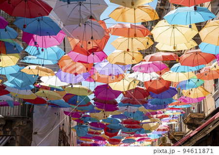 Catalina, Sicily, Italy. August 26, 2022. Multi colored umbrellas hanging amidst buildings with sky in background. View of decorative art on streets in residential district during summer. 94611871