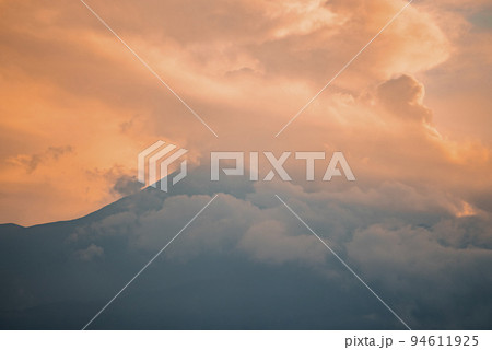 Scenic view of Mount Etna covered with smoke. Vibrant clouds wrapped around peak at twilight. Picturesque scenery of famous tourist attraction with orange sky in background during sunset. 94611925