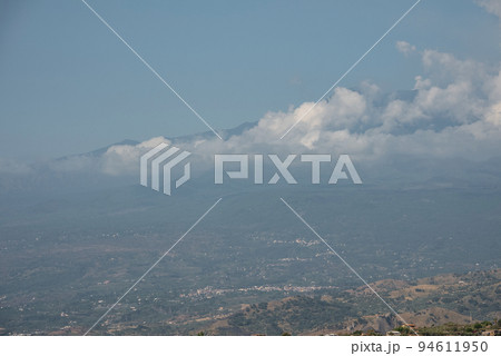 Beautiful landscape overlooking famous Mount Etna. Clouds covering active volcanic mountain with blue sky in background. Scenic view of tourist attraction during summer. 94611950