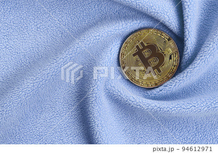 The golden bitcoin lies on a blanket made of soft and fluffy light blue fleece fabric with a large number of relief folds. The shape of the folds resembles a fan from a video card cooler The golden bitcoin lies on a blanket made of soft and fluffy light blue fleece fabric with a large number of relief folds. The shape of the folds resembles a fan from a video card cooler 94612971