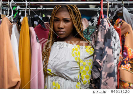 The serious face of the dark-skinned girl standing among the hanging clothes. 94613120