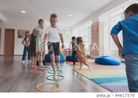 Small nursery school children with female teacher on floor indoors in classroom, doing exercise. Jumping over hula hoop circles track on the floor. Small nursery school children with female teacher on floor indoors in classroom, doing exercise. Jumping over hula hoop circles track on the floor. 94617570