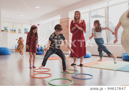 Small nursery school children with female teacher on floor indoors in classroom, doing exercise. Jumping over hula hoop circles track on the floor. Small nursery school children with female teacher on floor indoors in classroom, doing exercise. Jumping over hula hoop circles track on the floor. 94617647