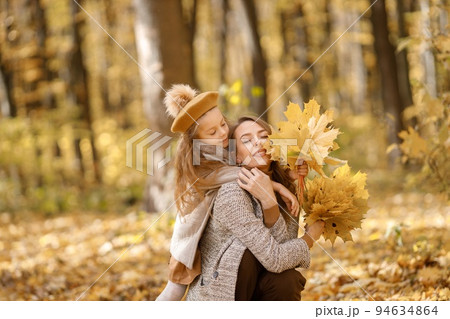 Young woman and little girl in autumn forest. Woman hugging her daughter. Girl wearing fashion clothes and holding yellow leaves. 94634864