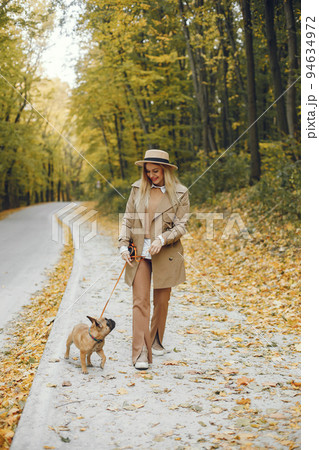 Woman and dog play and have fun in the autumn park. Brown french bulldog with female owner spend a day at the park walking. Woman wearing beige coat and a hat. Woman and dog play and have fun in the autumn park. Brown french bulldog with female owner spend a day at the park walking. Woman wearing beige coat and a hat. 94634972
