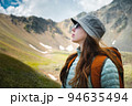 young woman tourist with a backpack and glasses stands in profile, summer portrait, against the backdrop of a mountain range 94635494