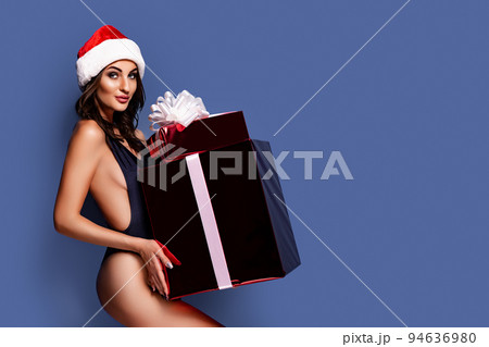 Portrait of young, sexy and beautiful woman in swimsuit and Santa hat, holding gift box. Girl in underwear lingerie. Blue background. Christmas, xmas, x-mas and winter concept Portrait of young, sexy and beautiful woman in swimsuit and Santa hat, holding gift box. Girl in underwear lingerie. Blue background. Christmas, xmas, x-mas and winter concept 94636980