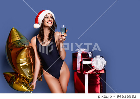Portrait of young, sexy and beautiful woman in swimsuit and Santa hat, holding golden baloon, glass of champagne near gift boxes. Christmas, xmas, x-mas and winter concept 94636982