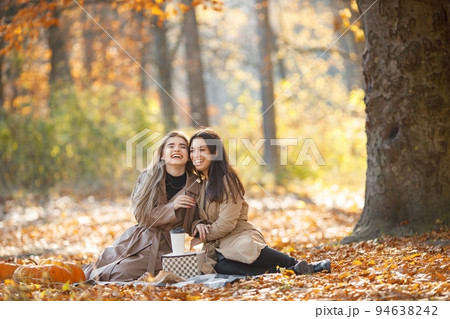 Two beautiful female friends spending time on a picnic blanket on the grass. Two young smiling sisters making picnic eating croissant in autumn park. Brunette and blonde girls wearing coats. 94638242