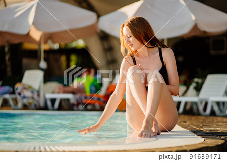 a slender young woman in a black swimsuit sits on the side of the pool. a slender young woman in a black swimsuit sits on the side of the pool. 94639471