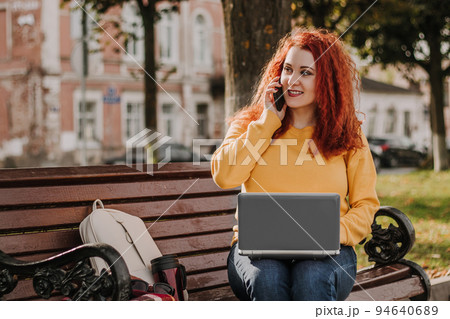 Young red-haired woman freelancer works in park using laptop. Modern businesswoman talking on phone. Online work on Internet. Young red-haired woman freelancer works in park using laptop. Modern businesswoman talking on phone. Online work on Internet. 94640689
