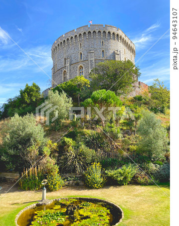 イギリス王室の公邸である石造りのウィンザー城、緑が生い茂るラウンドタワーの外観と青空と芝生 94643107