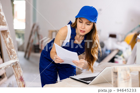 Female contractor in blue uniform using laptop at construction site Female contractor in blue uniform using laptop at construction site 94645233