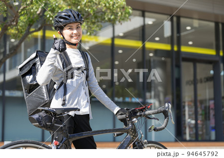 ロードバイクを前にしてデリバリーバックを背負って親指立てているフードデリバリー配達員の女性 ロードバイクを前にしてデリバリーバックを背負って親指立てているフードデリバリー配達員の女性 94645792