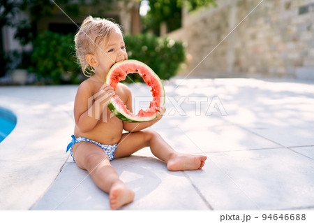Little girl gnawing a hole in a round piece of watermelon while sitting on a tile near the fence. High quality photo 94646688