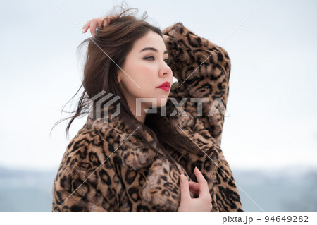 Winter portrait of brunette young woman with long hair straightens hair with his hand and looking away. Elegant authentic female dressed in faux fur coat with leopard pattern. 94649282