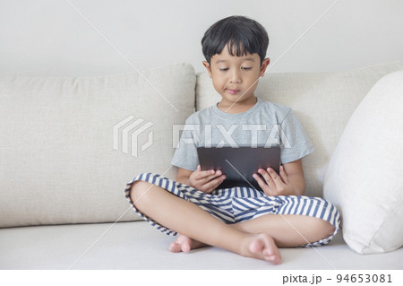 Adorable happy Asian boy wearing a gray shirt and blue-white striped shorts is having fun playing with his tablet on a cream sofa. looking at the mobile screen. Adorable happy Asian boy wearing a gray shirt and blue-white striped shorts is having fun playing with his tablet on a cream sofa. looking at the mobile screen. 94653081