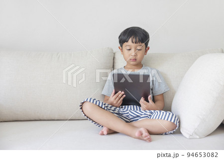 Adorable happy Asian boy wearing a gray shirt and blue-white striped shorts is having fun playing with his tablet on a cream sofa. looking at the mobile screen. Adorable happy Asian boy wearing a gray shirt and blue-white striped shorts is having fun playing with his tablet on a cream sofa. looking at the mobile screen. 94653082