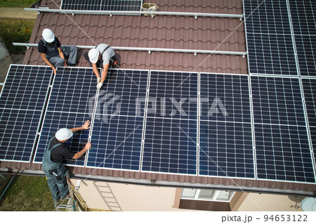 Men technicians mounting photovoltaic solar moduls on roof of house. Electricians in helmets installing solar panel system outdoors. Concept of alternative and renewable energy. Aerial view. Men technicians mounting photovoltaic solar moduls on roof of house. Electricians in helmets installing solar panel system outdoors. Concept of alternative and renewable energy. Aerial view. 94653122