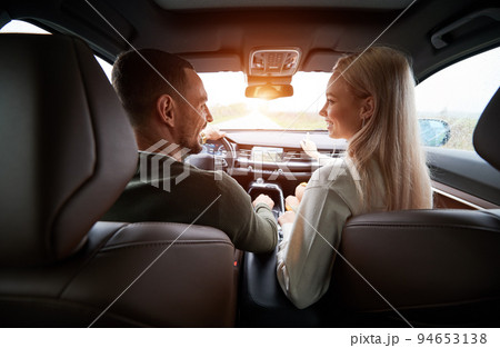 Back view of happy two people inside comfortable car. Joyful driver and cheerful female blonde in the front seat, sincerely smiling and looking at each other. Pleasant atmosphere during auto trip. Back view of happy two people inside comfortable car. Joyful driver and cheerful female blonde in the front seat, sincerely smiling and looking at each other. Pleasant atmosphere during auto trip. 94653138